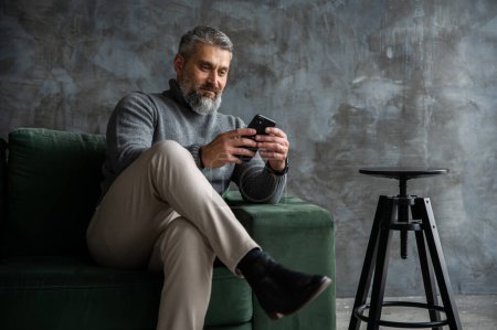 Téléchargez les photos : Homme sénior beau assis sur le canapé vert dans le salon loft, travaillant de la maison sur tablette. Designer masculin créatif préparant le projet seul - en image libre de droit