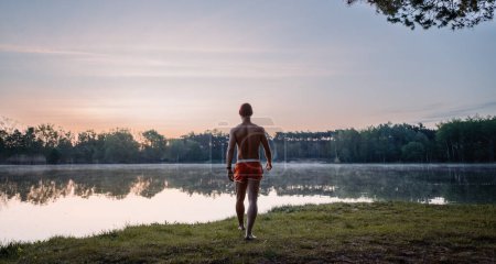 Téléchargez les photos : Back view of the good looking man in swim shorts going to the lake through the grass and looking at the water while preparing for the swimming in the winter morning. Ice swimming concept - en image libre de droit