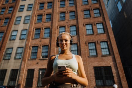 Téléchargez les photos : Jeune femme blonde sportive en tenue de fitness portant des écouteurs et tournant la musique sur smartphone avant de courir à l'extérieur. Concept de mode de vie sain - en image libre de droit