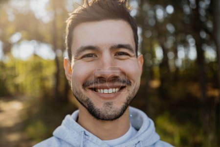 Téléchargez les photos : Portrait de jeune homme séduisant travaillant, souriant et posant dans la caméra en forêt, vêtu de vêtements de sport. - en image libre de droit