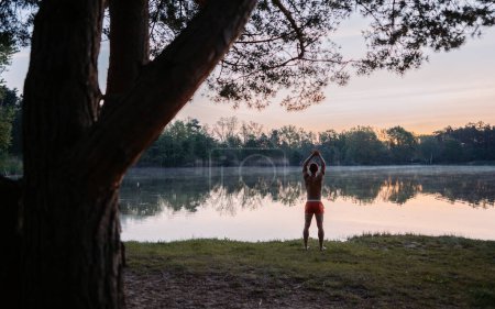 Téléchargez les photos : Portrait d'un bel homme s'étend avant de nager dans une rivière d'automne fraîche au lever du soleil le matin par les bois - en image libre de droit