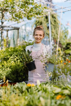 Téléchargez les photos : Vue verticale de la belle femme souriante jardinage et de prendre soin des plantes dans le centre de jardin. Poser dans la caméra - en image libre de droit