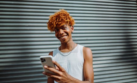 Téléchargez les photos : Portrait de l'adolescent multiracial homme souriant confiant tout en faisant selfie par la caméra à la rue. Un type maquillé avec une caméra de smartphone à l'extérieur dans la rue de la ville. Personnes et photographie - en image libre de droit