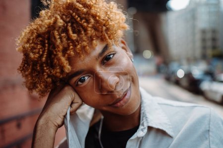 Téléchargez les photos : Portrait de jeune homme afro-américain attrayant posant et souriant dans la caméra, profitant de la vie, se maquillant dans la ville - en image libre de droit
