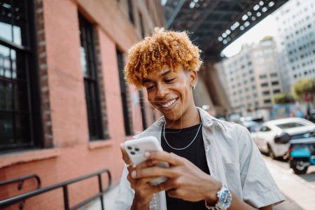 Téléchargez les photos : Portrait de jeune homme multiracial souriant heureux à l'aide d'un smartphone tout en marchant dans la ville. Adolescent bouclé profiter week-end tout en allant seul dans la rue vide - en image libre de droit