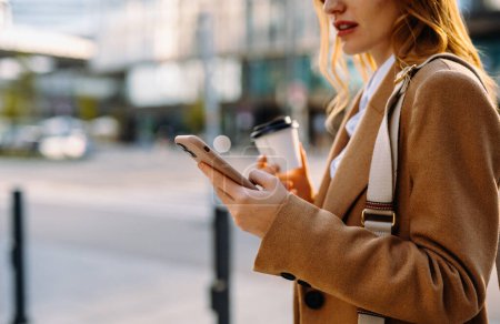 Téléchargez les photos : Focus élégante femme d'affaires en utilisant le téléphone, tenant le café avec des immeubles de condo résidentiels de ville floue en arrière-plan comme mode de vie progressif - en image libre de droit