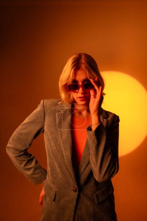 Téléchargez les photos : Portrait à la mode de dame blonde en lunettes de soleil posant isolée sur fond orange. Dame en tenue de veste à la mode gestuelle mains à la caméra - en image libre de droit