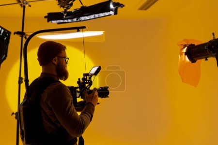Téléchargez les photos : Un vidéaste capte des images dans une pièce jaune à l'aide d'une caméra vidéo, mettant en valeur son talent dans l'industrie cinématographique en tant que cinéaste - en image libre de droit