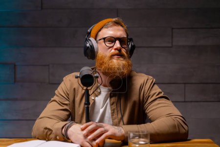 Téléchargez les photos : Un homme au visage barbu, portant lunettes et écouteurs, s'assoit à une table avec un microphone devant lui, prêt à enregistrer. Concept de studio Podcast - en image libre de droit