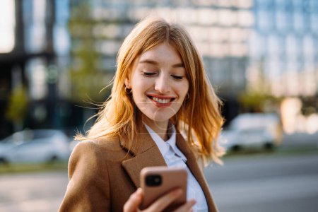 Téléchargez les photos : Une femme blonde heureuse aux cheveux superposés sourit en regardant son téléphone portable, un appareil de communication portable. Son geste joyeux montre qu'elle utilise un gadget en voyage - en image libre de droit