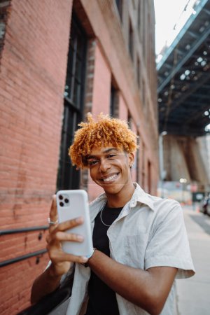 Téléchargez les photos : Un jeune homme vêtu de vêtements vintage sourit en prenant un selfie avec son téléphone portable dans la rue, présentant ses lunettes et ses manches retroussées. - en image libre de droit