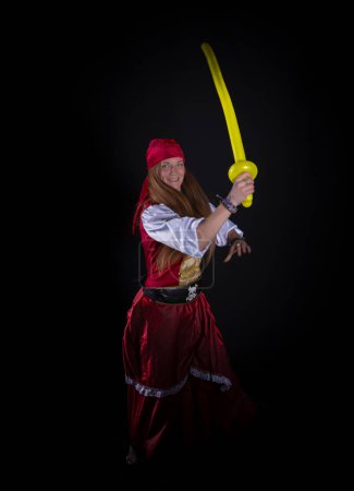 Foto de Woman with long hair dressed as a pirate in a red suit and with a yellow balloon in the shape of a sword in her hand and on a black background photography of party events - Imagen libre de derechos