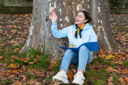 Téléchargez les photos : Une fille orientale est assise sous un arbre avec un dossier bleu dans les mains et rit, Autriche. Photo de haute qualité - en image libre de droit