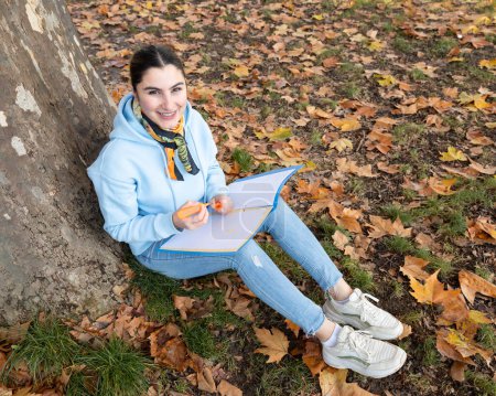 Téléchargez les photos : Une étudiante s'assoit sous un arbre avec un dossier bleu à la main et sourit, Autriche, Salzbourg. Photo de haute qualité - en image libre de droit