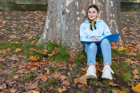 Téléchargez les photos : Oriental girl assis sous un arbre avec un dossier bleu dans ses mains, Autriche. Photo de haute qualité - en image libre de droit