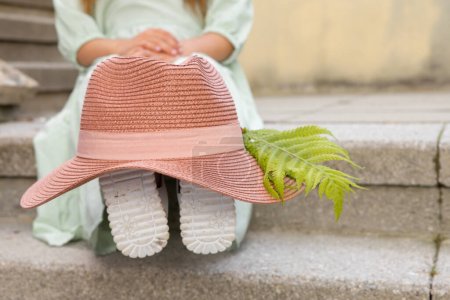 Téléchargez les photos : Une fille en robe romantique est assise sur les marches en pierre, portant un chapeau rose avec une plume de fougère sur les jambes. Photo de haute qualité - en image libre de droit