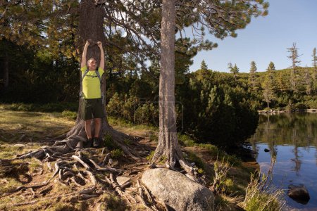 Foto de Un hombre con una mochila está parado cerca de un árbol en la orilla de un lago de montaña, levantando sus manos, Austria. Foto de alta calidad - Imagen libre de derechos