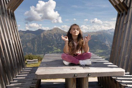 Téléchargez les photos : Petite fille mignonne aux cheveux longs assise sur une table en bois dans un belvédère en position lotus sur le fond des montagnes et du ciel. Photo de haute qualité - en image libre de droit