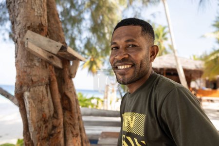 A smiling middle-aged Papuan sits against the backdrop of a Papuan village and the ocean. High quality photo