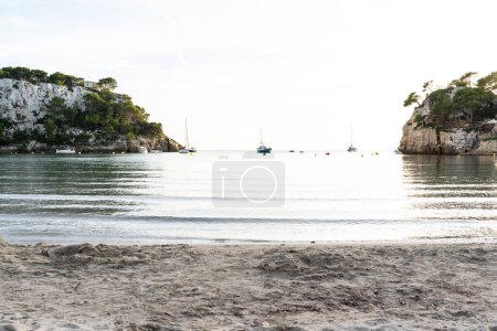 Foto de Atardecer en una playa de arena con acantilados y embarcación al fondo en la Cala Galdana situada en Menorca, una isla de España. - Imagen libre de derechos