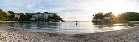 Photo for Panoramic view of Cala Galdana in Minorca at sunset, a sand beach with cliffs, vessel on the background and a couple in silhouette in the middle. - Royalty Free Image