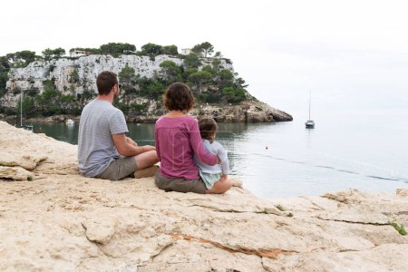 Foto de Vista trasera de una pareja con su hijita sentada al borde de un acantilado mirando vistas en Cala Galdana, Menorca. - Imagen libre de derechos