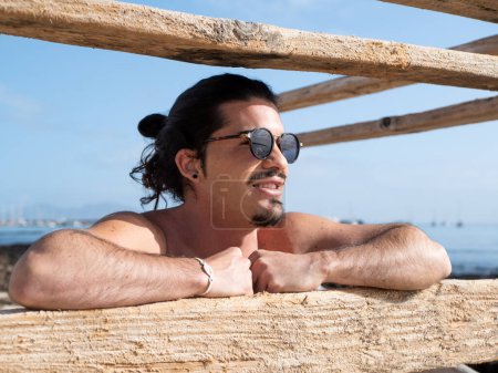 Foto de Hombre hispano sin camisa con gafas de sol retrato tomando el sol y apoyándose en una valla junto al mar - Imagen libre de derechos