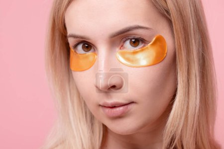 Portrait of pretty girl applying golden collagen patches under her eyes in unusual way. Woman facial treatment . Cosmetology, beauty and skincare around eyes, preventing wrinkles.