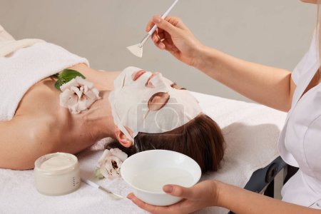 esteticista alisa la máscara de la hoja en la cara de la mujer para rehidratar la piel de la cara, anti arrugas procedimiento cosmético en salón de spa de belleza. Cosmetólogo aplicando mascarilla humectante en la cara femenina