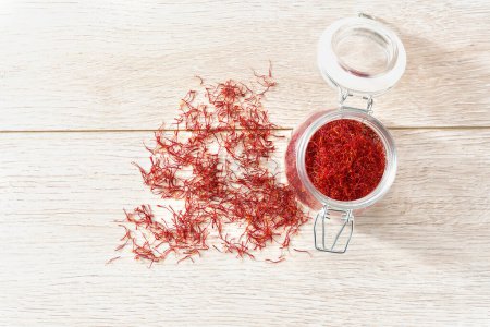 Photo for Open storage jar with saffron on a wooden table, top view. Saffron stigmas scattered on a wooden surface from a glass bottle. - Royalty Free Image