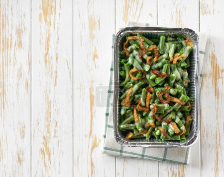 Téléchargez les photos : Boîte à nourriture en aluminium avec casserole de haricots verts avec oignons frits croustillants sur une table blanche, vue sur le dessus. Boîte à nourriture à feuilles avec repas à emporter. - en image libre de droit