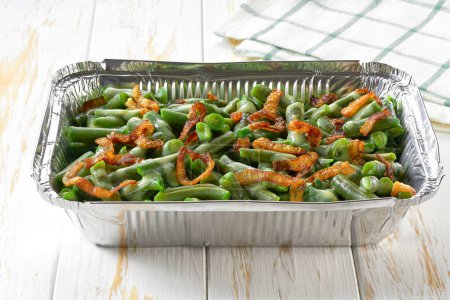 Photo for Green bean casserole with crispy fried shallots onion in a disposable square aluminium foil baking dish on a white table. Foil food container tray. - Royalty Free Image