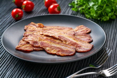 Téléchargez les photos : Bacon frit avec sauce barbecue dans un plat sur une table noire. Morceaux de bacon sautés croquants frits dans une assiette noire. - en image libre de droit