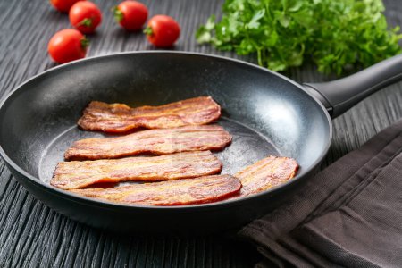 Téléchargez les photos : Lanières de bacon frit dans une poêle sur une table de cuisine noire. Étourdissant morceaux de bacon chaud dans une poêle. Idée petit déjeuner. - en image libre de droit