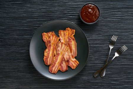 Téléchargez les photos : Assiette avec bacon frit et sauce barbecue sur une table, copier l'espace pour le texte. Assiette avec bacon frit sur table de cuisine noire. - en image libre de droit