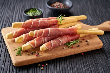 Photo for Grissini-traditional italian bread snack with Parma ham prosciutto and rosemary on a black wooden table . - Royalty Free Image