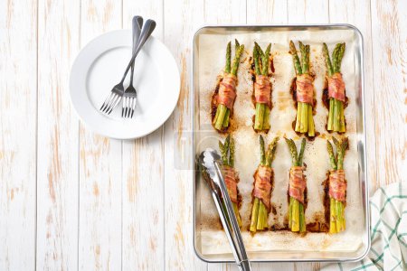 Photo for Roasted bundles of green asparagus wrapped in a bacon on a baking tray on wooden table with copy space for text.Top view. - Royalty Free Image