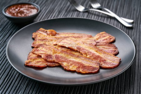 Téléchargez les photos : Assiette avec bacon frit et sauce barbecue sur une table noire. Assiette avec bacon frit sur table de cuisine noire. - en image libre de droit