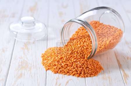 Photo for Organic red lentils spill out of a glass storage jar on a light table, selective focus. - Royalty Free Image