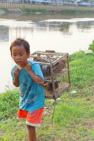 Téléchargez les photos : Jakarta, Indonésie - 13 mars 2023 : Un garçon passe près de la rivière tout en tenant une cage en bois remplie de pigeons dans l'après-midi. - en image libre de droit