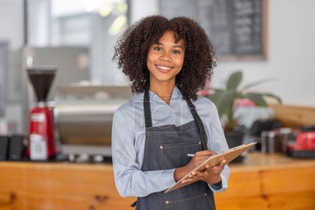 Eine afrikanische Coffeeshop-Inhaberin mit Schürze steht vor dem Schalter und führt einen Aktiencheck durch. Afro-Haar Mitarbeiter Barista Unternehmer.
