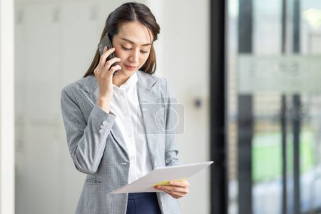 Téléchargez les photos : Jeune femme asiatique souriante d'affaires parlant sur le téléphone mobile, debout dans le bureau de travail avec le document dans sa main - en image libre de droit