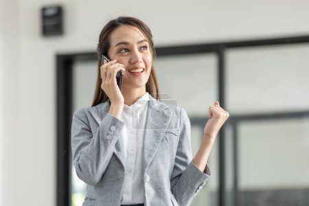 Téléchargez les photos : Jeune souriant heureux excité entreprise asiatique femme parler sur téléphone mobile, debout dans le bureau de travail avec document dans sa main - en image libre de droit