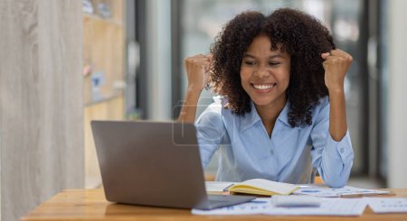 Téléchargez les photos : Heureuse femme africaine asiatique excitée regardant l'écran de l'ordinateur portable, célébrant une victoire en ligne, ravie jeune fille africaine criant de joie, isolée sur un fond de bureau - en image libre de droit