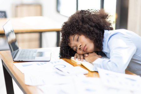 Foto de Mujer africana agotado milenario durmiendo en su escritorio de la oficina, al lado de la computadora portátil y documentos, cansado de exceso de trabajo. Niña africana joven adicta al trabajo que sufre de fatiga crónica en el lugar de trabajo - Imagen libre de derechos