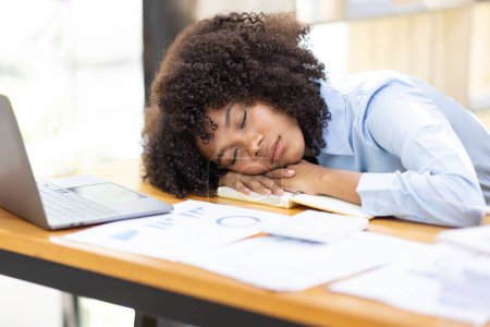Erschöpfte Afrikanerin, die erschöpft auf ihrem Bürotisch neben Laptop und Dokumenten schläft, müde von Überarbeitung. Junge afrikanische Workaholic-Mädchen leiden unter chronischer Müdigkeit am Arbeitsplatz