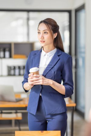 Téléchargez les photos : Portrait de jeune femme asiatique hoding tasse de café et debout dans un lieu de travail moderne occupé - en image libre de droit