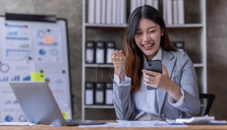 Téléchargez les photos : Heureux heureux heureux succès asiatique femme d'affaires triomphant avec smartphone et ordinateur portable sur le lieu de travail. - en image libre de droit
