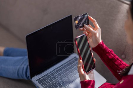 Joven mujer asiática bastante segura de trabajar con el ordenador portátil y la tarjeta de crédito en el sofá en casa, compras y pago en línea mediante el uso de ordenador portátil con efecto de luz solar.