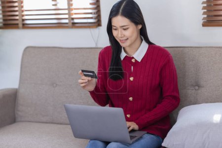 Téléchargez les photos : Jeune confiante jolie femme asiatique travaillant avec ordinateur portable et carte de crédit sur canapé à la maison, les achats et le paiement en ligne en utilisant un ordinateur portable avec effet de lumière du soleil. - en image libre de droit